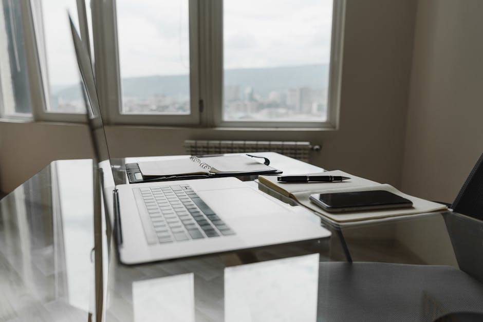 A modern office setup featuring a laptop, notebook, and pen on a glass table with city view.